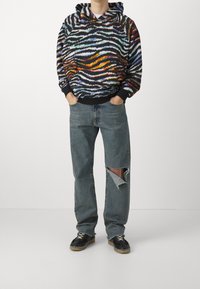 Sudadera con capucha con patrón de rayas de cebra multicolor, puños negros; combinada con jeans azul desgastados con un gran desgarrón y zapatillas negras.