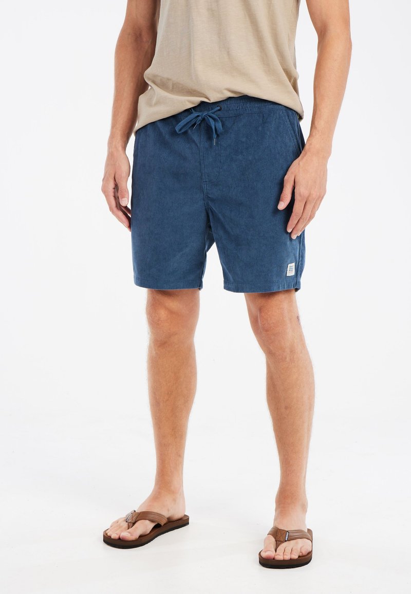 Marineblauwe zwemshorts van een gestructureerde stof, met elastische tailleband, koord en vo pockets. Gemaakt in combinatie met bruine slippers.