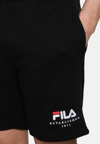 Los pantalones cortos de algodón negros cuentan con una cintura elástica y bolsillos laterales. El dobladillo izquierdo presenta un logotipo bordado en rojo, blanco y negro.