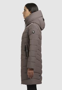 Manteau long en duvet gris, avec un col haut, un design matelassé horizontal, des poches latérales et zippées, ainsi qu'un logo brodé sur la manche.