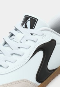 Sneaker in pelle bianca con inserti in suede grigio e logo in gomma nera. Presenta lacci piatti, pannello laterale testurizzato e suola in gomma color caramello.