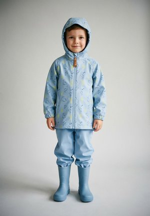 Young child wearing light blue hooded raincoat with duck prints, matching rain pants, and blue rubber boots, standing against plain background.