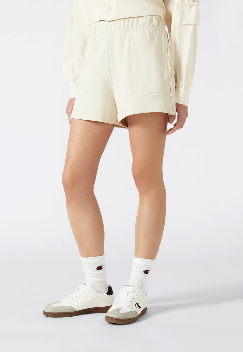 Crème-kleurige shorts van zacht materiaal met een elastische tailleband en twee voorzakken, gecombineerd met witte sneakers en sokken met een logo.