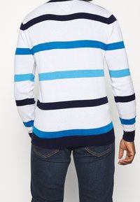 Stickad tröja i vitt med djärva horisontella ränder i marinblått, teal och ljusblått. Designen inkluderar en V-ringning och långa ärmar.