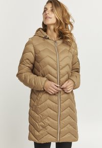 Pufferjakke i en varm beige farve med et chevronmønster, hætte og lynlås foran. Designet inkluderer elastiske manchetter og sidelommer.