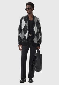 Cardigan argyle noir et gris, coupe oversize, fabriqué en mélange de laine douce. Porté avec un pantalon noir, des lunettes de soleil élégantes et un sac noir.