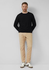 Mann mit kurzen Haaren, der ein schwarzes langärmliges Shirt, beige Hosen und weiße Sneaker mit schwarzen Akzenten trägt, steht vor einer schlichten grauen Wand.