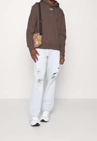 Sudadera marrón con logo en el frente, vaqueros desgastados de color azul claro, bolso bandolera beige y zapatillas blancas con detalles negros.