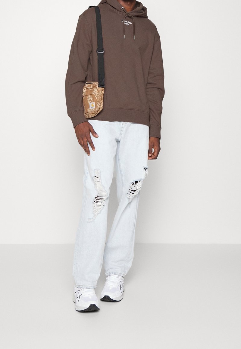 Sudadera marrón con logo en el frente, vaqueros desgastados de color azul claro, bolso bandolera beige y zapatillas blancas con detalles negros.