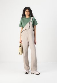 Beige salopette met een diagonale bies en verstelbare banden, gecombineerd met een groene T-shirt met korte mouwen. Draagt een kleine gele tas.