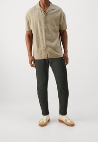 Beige kortermet skjorte med krave, knappet foran; parret med mørkegrønne bukser og cremefarvede sneakers med brune accenter.