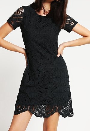 Kobieta w czarnej, krótkiej sukience szydełkowej z okrągłymi wzorami, stojąca z rękami na biodrach na jednolitym tle.