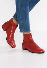 Rote Lederstiefeletten mit einem niedrigen Blockabsatz, seitlichem Reißverschluss und glatter Textur, kombiniert mit Jeans mit fransigem Saum auf einem weißen Hintergrund.