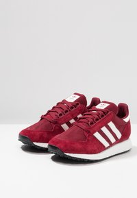 Röda mocka- och nätträningsskor med vita ränder, rund tå och texturerad gummisula. Funktioner inkluderar snörning och vadderad krage.