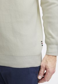 Light beige ribbed sweater with textured fabric, featuring a white, black, and red striped accent tag at the hem.