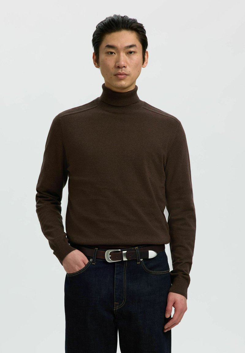 Homme portant un pull à col roulé marron foncé et un jean foncé avec une ceinture marron, debout avec une main dans la poche sur un fond uni.