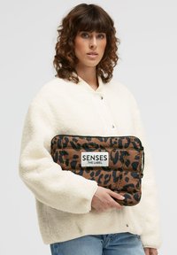 Braune gesteppte Clutch mit schwarzem Leopardenmuster, gehalten gegen eine weiße Fleecejacke. Verfügt über ein weißes Etikett mit dem Text "SENSES .THE LABEL".