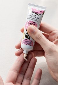 Korres Damask Rose-masktub, vit med rosa och gyllene blommönster, som fördelar en krämig, off-white produkt på en hand.