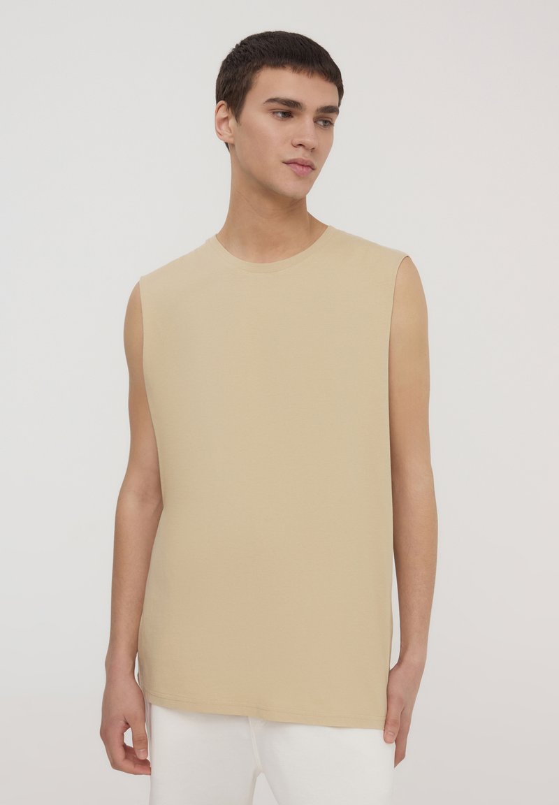 Camicetta beige senza maniche realizzata in morbido cotone, caratterizzata da un collo rotondo e una vestibilità rilassata. Texture liscia con orlo dritto, senza motivi visibili.