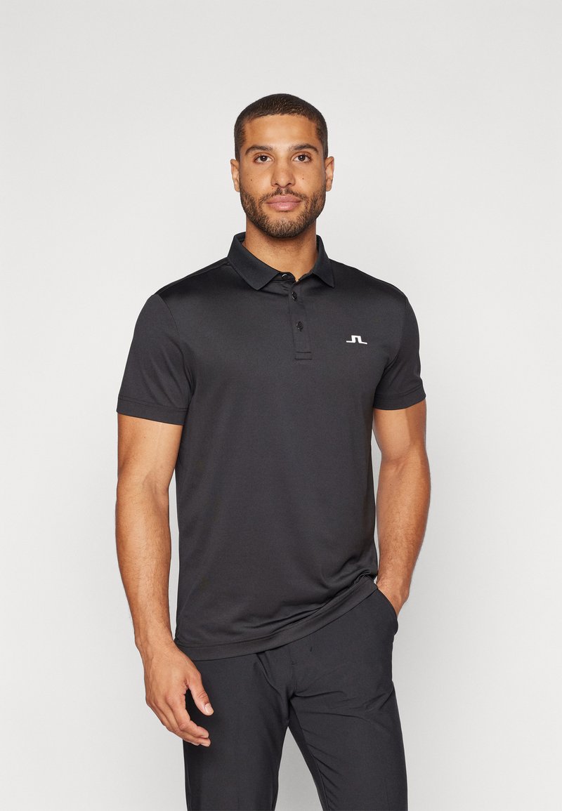 Polo nero in tessuto morbido con colletto, maniche corte e un piccolo logo bianco sul lato sinistro del petto; design aderente, abbinato a pantaloni scuri.