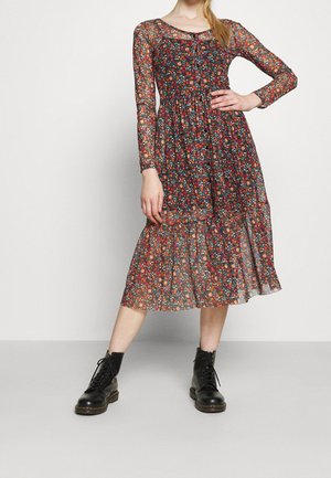 Vrouw die een doorschijnende midi-jurk met lange mouwen en bloemenprint draagt, gecombineerd met zwarte vetercombatlaarzen, staand met één hand op de heup en de benen uit elkaar.