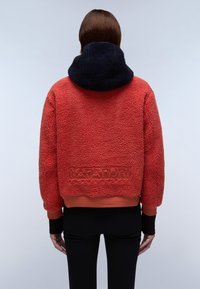 Rød shearling jakke med en marineblå hætte, cropped design og ribbede manchetter. Har prægede logo på ryggen og en blød tekstur.