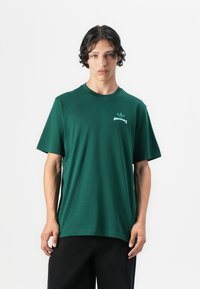 Zöld pamut póló kerek nyakkal, rövid ujjú, a mellen apró fehér Adidas logóval. Sima textúra, laza szabás.