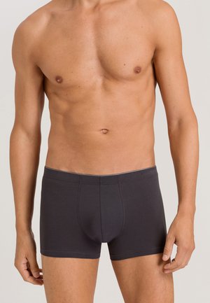 Oberkörper eines oberkörperfreien Mannes, der dunkelgraue Boxershorts trägt, vor einem schlichten hellen Hintergrund, Arme entspannt an den Seiten.
