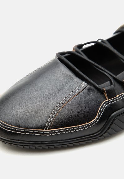 Κοντινή λήψη του μπροστινού μέρους μαύρου δερμάτινου παπουτσιού με μπεζ ραφές, ελαστικά κορδόνια και υφασμένη ελαστική σόλα σε λευκό φόντο.