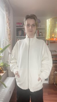 Bílá zipová bunda s vysokým límcem, dlouhými rukávy a bočními kapsami. Měkká textilie, volný střih, minimální značení na hrudi.