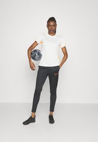 Valge spordipaita, millel on oranžid aktsendid, koos mustade kitsaste pükstega, millel on väike oranž logo, ning mustade spordijalatsitega.