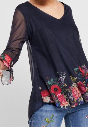 Femme porte un top bleu marine transparent avec un imprimé floral coloré en bordure et un jean brodé avec des motifs floraux sur la cuisse.