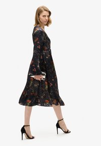 Vestido negro con flores, mangas largas acampanadas y falda en capas, con patrones rojos y verdes. Acompañado de sandalias de tacón alto negras.