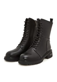 Botas negras hasta el tobillo con un acabado de cuero liso, cordones en la parte frontal, cremallera lateral y suela gruesa. Cuenta con detalles decorativos en la parte superior.