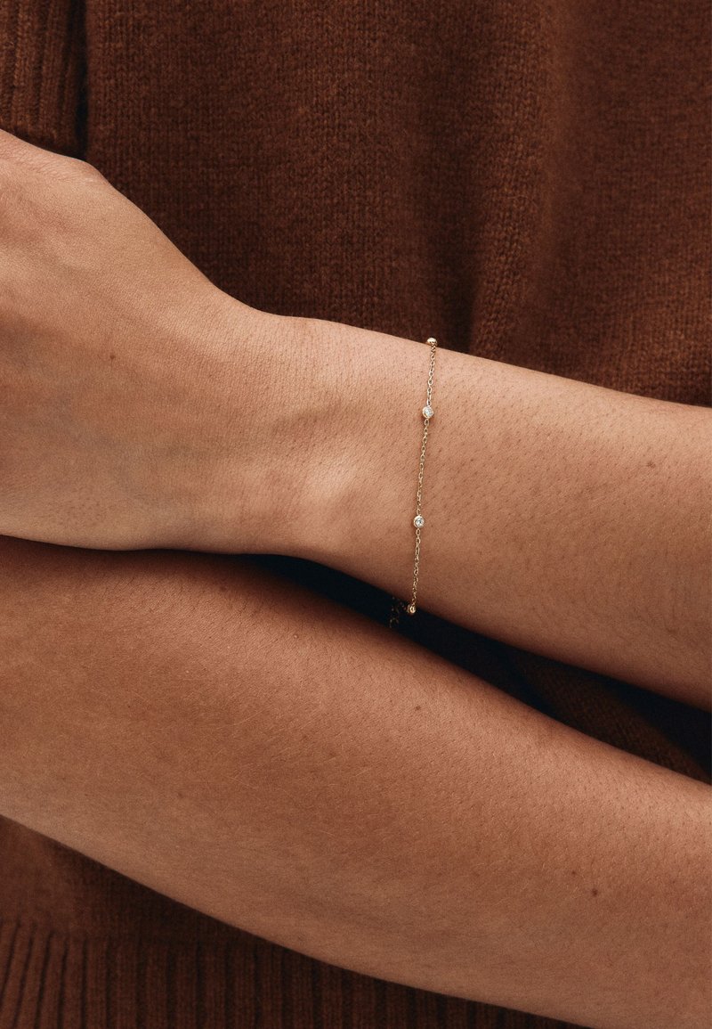 Auksinė grandinėlės apyrankė su mažais apvaliais akcentais ir subtiliu dizainu, dėvima ant riešo priešais rudą megztinio foną.