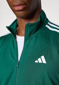 Zaļa sportiska zip jaka, kas izgatavota no gludas auduma, ar baltām svītrām uz pleciem un logo apakšējā labajā stūrī.