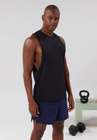 Camisola desportiva preta sem mangas com cavas alongadas, combinada com calções azuis marinhos. Tecido liso, design atlético, vestida por um modelo masculino.