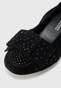 Μαύρο φλατ παπούτσι από σουέντ με ευρεία φιόγκο διακοσμημένη με λαμπερά μαύρα τρουκς. Στρογγυλερή μύτη με ομαλή υφή.
