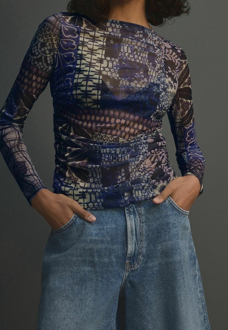 Personne portant un haut ajusté à manches longues avec des motifs bleus et violets et un jean en denim bleu clair ample, les mains dans les poches.