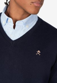 Maglione blu navy a collo a V in morbido tessuto, indossato sopra una camicia a bottoni azzurri. Logo ricamato evidente in colore contrastante sul petto.