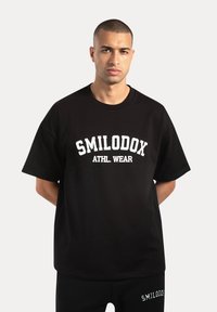 Camiseta de algodón negra con la inscripción "SMILODOX ATHL. WEAR" en letras blancas en negrita, de manga corta, corte relajado y cuello redondo.