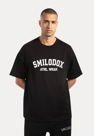 Camiseta de algodón negra con la inscripción "SMILODOX ATHL. WEAR" en letras blancas en negrita, de manga corta, corte relajado y cuello redondo.