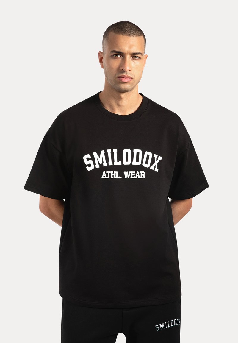 Camiseta de algodón negra con la inscripción "SMILODOX ATHL. WEAR" en letras blancas en negrita, de manga corta, corte relajado y cuello redondo.