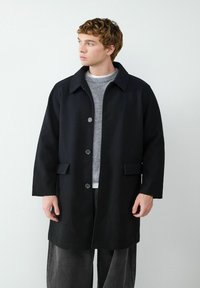 Schwarzer Wollmischmantel mit Kragen, sechs Knöpfen und zwei vorderen Taschen, getragen über einem grauen Pullover und schwarzen Hosen. Glatte Textur.