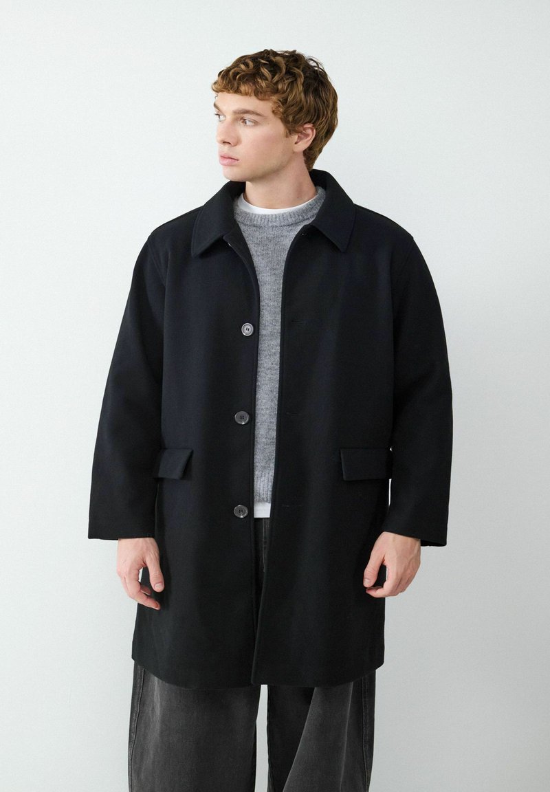 Schwarzer Wollmischmantel mit Kragen, sechs Knöpfen und zwei vorderen Taschen, getragen über einem grauen Pullover und schwarzen Hosen. Glatte Textur.