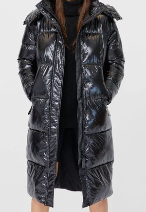 Long manteau matelassé noir brillant avec capuche, porté sur un pull à col roulé noir et une jupe, mains dans les poches.