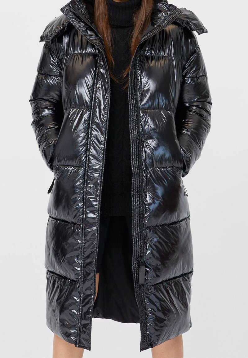 Long manteau matelassé noir brillant avec capuche, porté sur un pull à col roulé noir et une jupe, mains dans les poches.