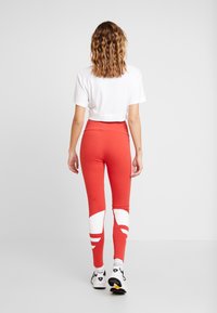 Červené legíny s bílými akcenty, vysoký pas, hladká textura a přiléhavý střih. Kombinované s krátkým bílým tričkem a sportovními botami.