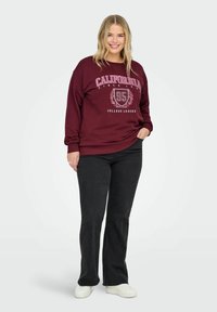 Burgundijas sporta krekls ar tekstu "CALIFORNIA" un grafiku, ribotām piedurknēm, brīva piegriezuma. Kombinēts ar melnām platām biksēm un baltām apavām.