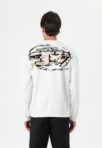 Sweatshirt branco com uma grande ilustração abstrata em rosa, amarelo e cinza nas costas. Apresenta mangas longas e punhos e bainha canelados.